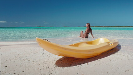 Woman relax sitting on beach, yellow kayak on sand, blue sky, turquoise water. Tanned young female on tropical island back view. Outdoor lifestyle travel on summer holiday vacation, active sport