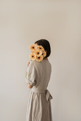 Beautiful woman in linen dress holding pastel gerber flowers bouquet over white wall. Aesthetic holiday celebration concept