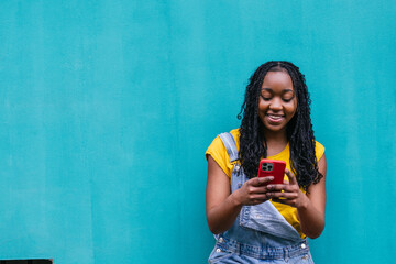 Smiling young Latinx woman in yellow shirt using a red smartphone, standing against a bright blue...