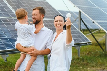 Solar energy concept. A young, happy family is standing near solar panels and holding an electric...