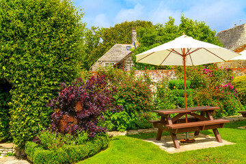 A picnic table with umbrella on a sunny day in an English garden outside a stone cottage.