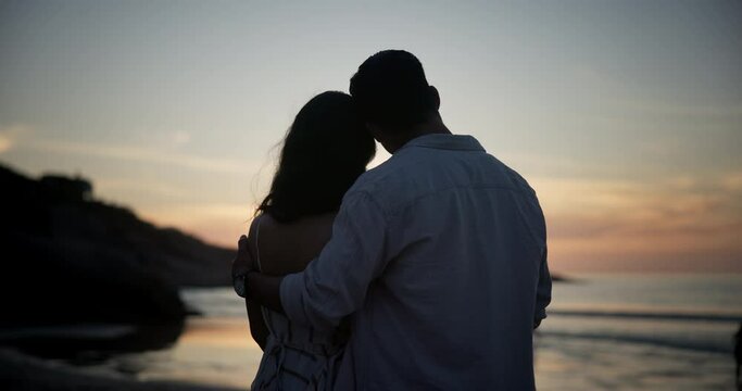 Sunset, back and couple at ocean for love on holiday, travel or summer vacation on valentines day date. Silhouette, man and woman at beach together for connection, hug and relationship at sea outdoor
