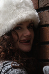 portrait of flirtatious cheerful beautiful young woman in fur winter hat leaning against brick wall