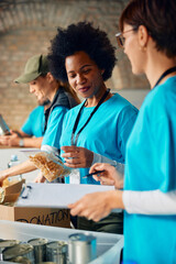 Smiling black woman and her coworkers packing food at volunteer center. - obrazy, fototapety, plakaty