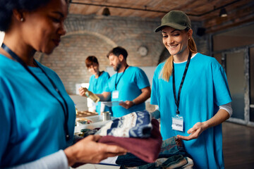 Happy volunteers packing donated clothes while working at humanitarian aid center. - obrazy, fototapety, plakaty