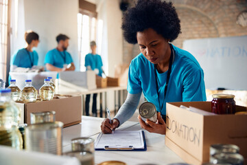 Black female volunteer making list of donated food at charitable foundation.