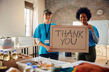 Happy female volunteers with 'Thank you' placard at donation center looking at camera. - obrazy, fototapety, plakaty