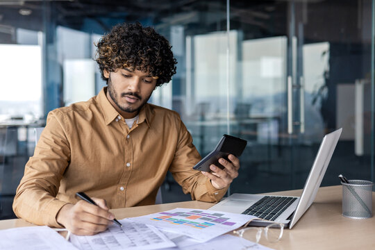 Serious and focused young Indian man working in office at desk with documents. Processes data and accounts, uses a notebook and a calculator.