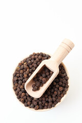 Black peppercorns in bowl with wooden scoop on white background. Dry white pepper grain