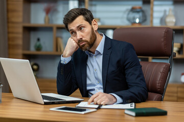 A young businessman man in a suit sits in the office at a desk in front of a laptop, is sad and...