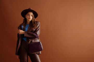 Fashionable confident woman wearing hat, faux leather suit blazer, pants, holding trendy purple...