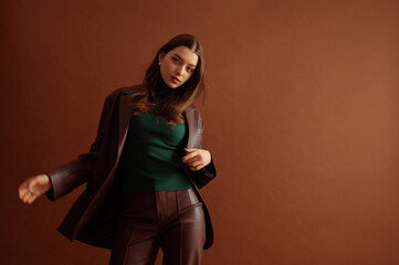 Fashionable confident woman wearing trendy faux leather suit blazer, pants, green ribbed turtleneck, posing on brown background. Studio fashion portrait. Copy, empty, blank space for text