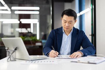 Fototapeta na wymiar Serious thinking man behind paper work inside office, mature asian man in business suit filling data in table, writing with pen, businessman boss at workplace with laptop.