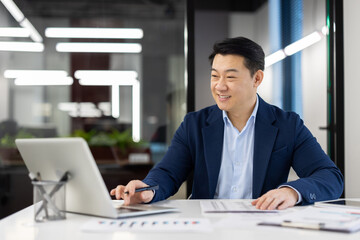 Satisfied with achievement male financier accountant inside office at workplace, Asian businessman smiling happy working with accounts contracts and reports, using laptop.