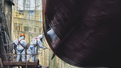 Shipyard Painters wearing personal protective clothing, gas masks and hazmat suits apply paint to...