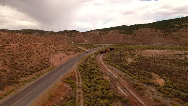 Aerial: Drone Forward Shot Of Railway And Car Moving In Semi Arid Desert With Hills Under Clouds - Ely, Nevada