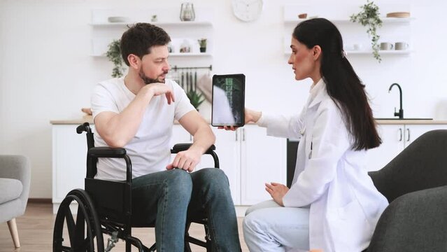 Adult Caucasian woman doctor checking up condition of mature bearded male patient at home. Young female nurse talking to mature man wheelchair user, showing x-ray scan of spine on modern tablet.