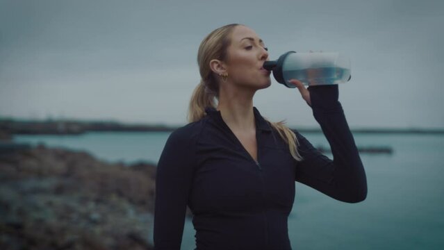 Female drinking protein shake, hydration exercising cardio workout training focused female athlete runner in cloudy seaside background tracking close up