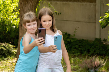 Two young girls, children smiling as they take a selfie with their smartphones mobile phones outdoors garden during daytime Kids and social media technology and internet concept photography, lifestyle