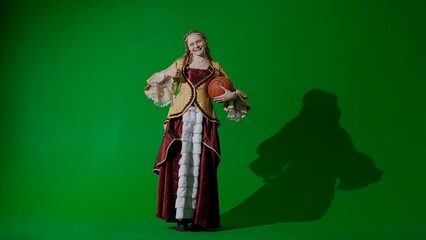 Woman in ancient outfit on the chroma key green screen background. Female in renaissance style dress holding basketball ball smiles shows thumb up.