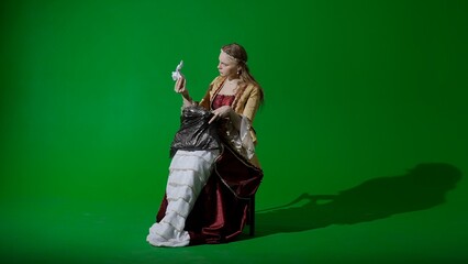 Woman in ancient outfit on the chroma key green screen background. Female in renaissance dress holding garbage bag looks inside takes out trash cans.