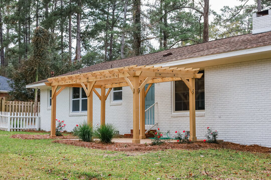 The rear back exterior of a newly painted and renovated white brick ranch style house with a large yard and a newly built wood arbor pergola for shade
