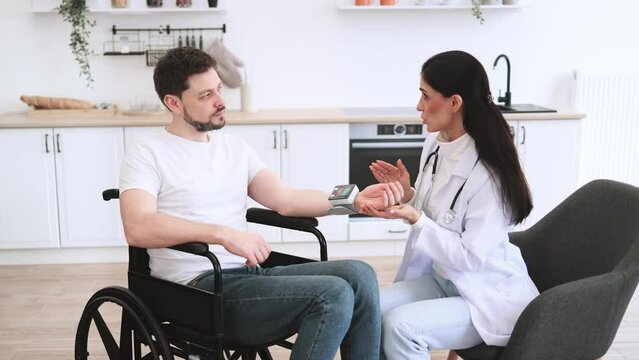 Adult man in wheelchair listening explanations about necessity of leading healthy lifestyle. Attractive cardiologist telling patient with disability about all reasons of hypertension sitting at home.