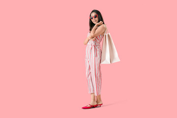 Happy beautiful young Asian woman in sunglasses with shopping bags on pink background