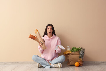 Shocked young woman with store receipts and food sitting near beige wall. Price rise concept