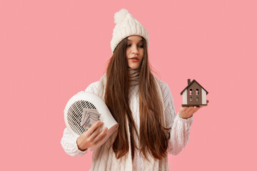 Young woman with electric fan heater, money and house model on pink background. Price rise concept