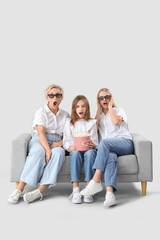 Fototapeta premium Shocked little girl with her mom and grandmother watching movie on sofa against light background