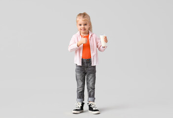 Cute little girl with glass of milk showing thumb-up on light background