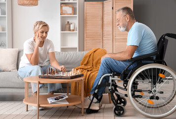 Mature woman with her husband in wheelchair playing chess at home