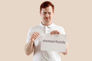 Young man holding paper with word HEMORRHOIDS and sad smile on beige background