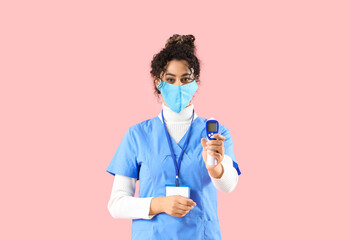 Female African-American medical intern in mask with infrared thermometer on pink background