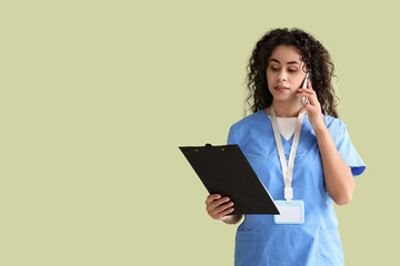 Female African-American medical intern with clipboard talking by mobile phone on green background