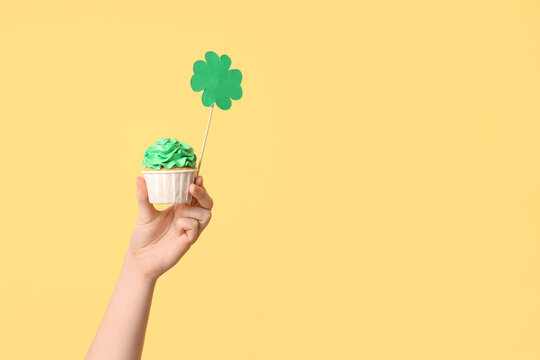 Female hand with tasty cupcake and paper clover for St. Patrick's Day on beige background