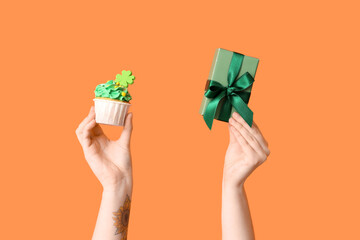 Female hands with tasty cupcake and gift box for St. Patrick's Day on orange background