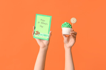 Fototapeta na wymiar Female hands with tasty cupcake and festive postcard for St. Patrick's Day on orange background