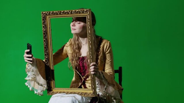 Woman in ancient outfit on chroma key green screen background. Female in renaissance style dress holding painting frame taking selfie on smartphone.