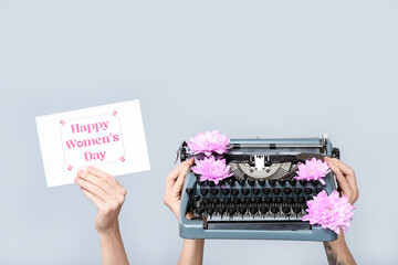 Hands holding vintage typewriter, flowers and festive postcard with text HAPPY WOMEN'S DAY on grey...