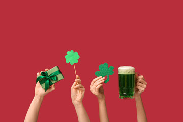 Female hands holding beer, gift box and decorative clovers on red background. St. Patrick's Day...