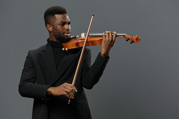 Elegant African American man in black suit playing violin on gray background in music performance setting