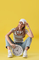 Young woman with clock sitting on yellow background