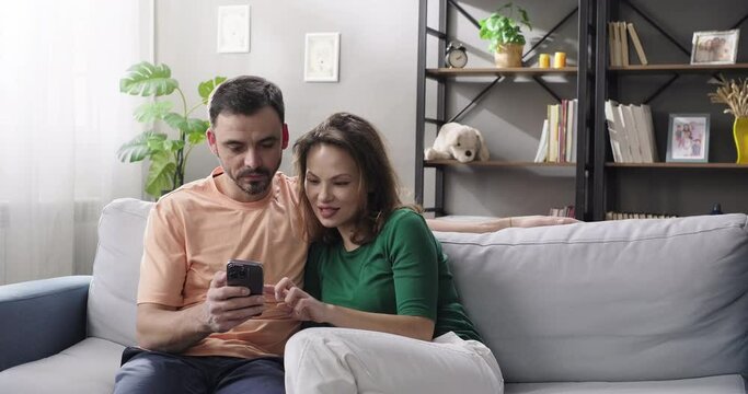 Couple using smartphone, shopping online, ordering delivery, checking social media apps, watching videos while sitting on couch in the living room
