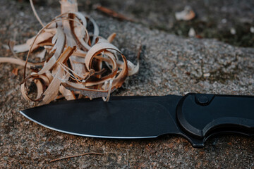 A black pocket knife sitting by some wood shreds to start a fire in the wilderness