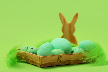 Wicker tray with painted Easter eggs, feathers and wooden bunny on green background