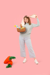 Beautiful young happy woman in bunny ears with carrot-shaped toys and Easter basket on pink...