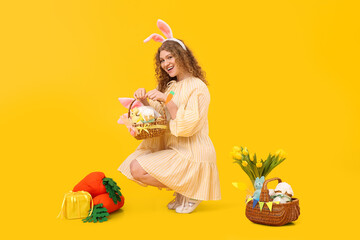 Beautiful young woman in bunny ears with Easter baskets, carrot-shaped toys and flowers on yellow background