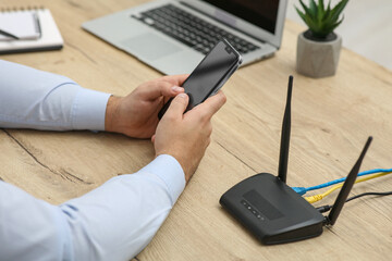 Man with smartphone and laptop connecting to internet via Wi-Fi router at wooden table, closeup